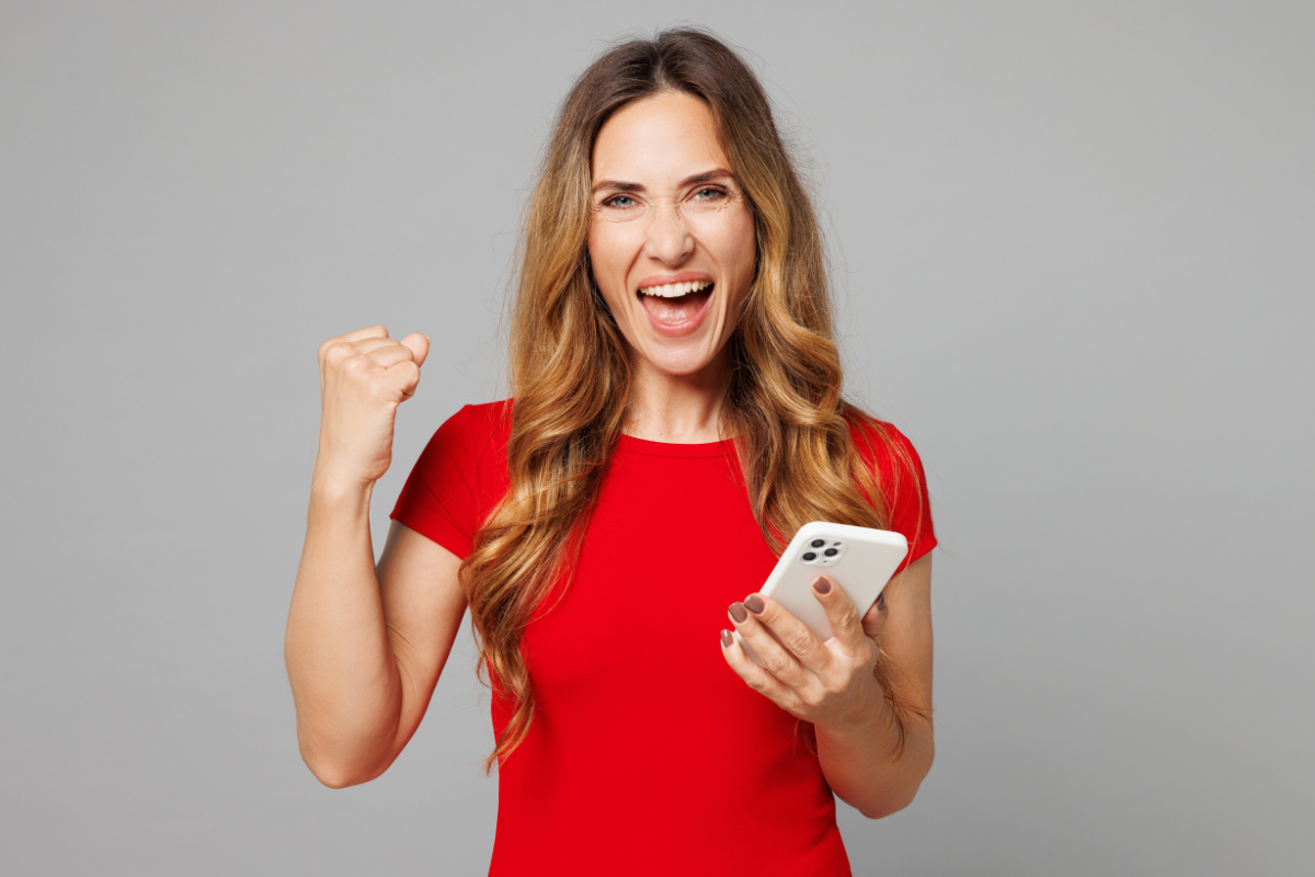Young excited overjoyed cool happy woman she wear red t-shirt casual clothes hold in hand use mobile cell phone do winner gesture isolated on plain grey background studio portrait. Lifestyle concept