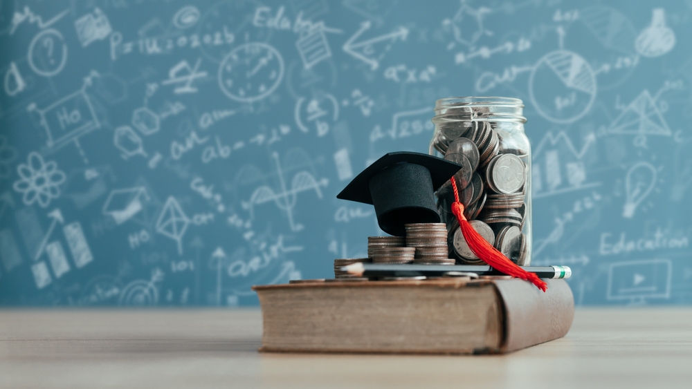 Graduation cap on top of stack of coins on top of book; jar of coins behind graduation cap