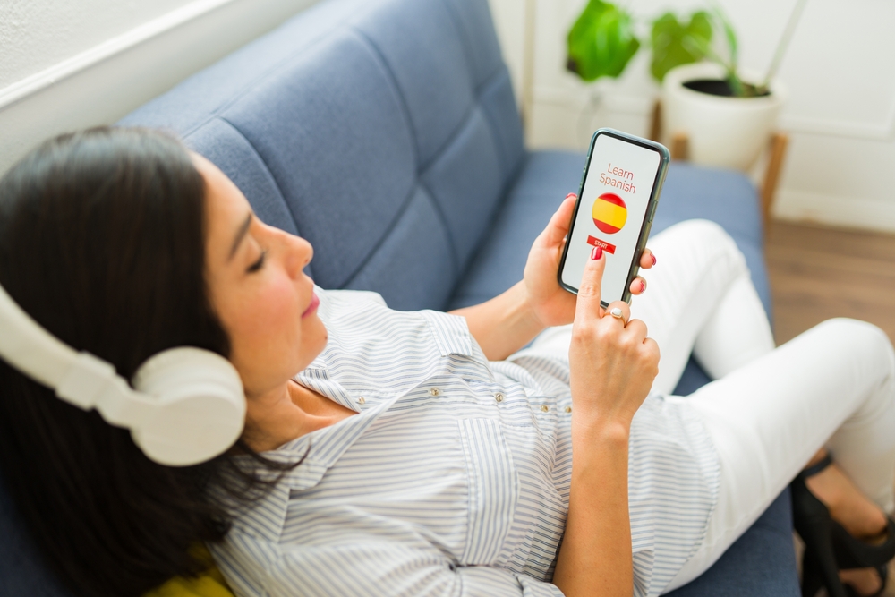 Woman wearing headphones while holding smartphone; "Learn Spanish" and "Start" button on smartphone screen