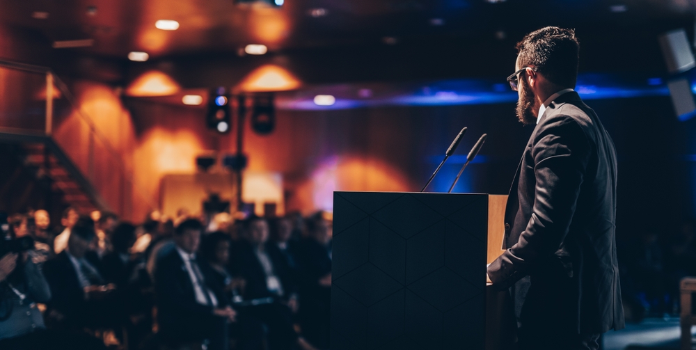 Speaker giving a talk at a conference; conference hall audience full of people