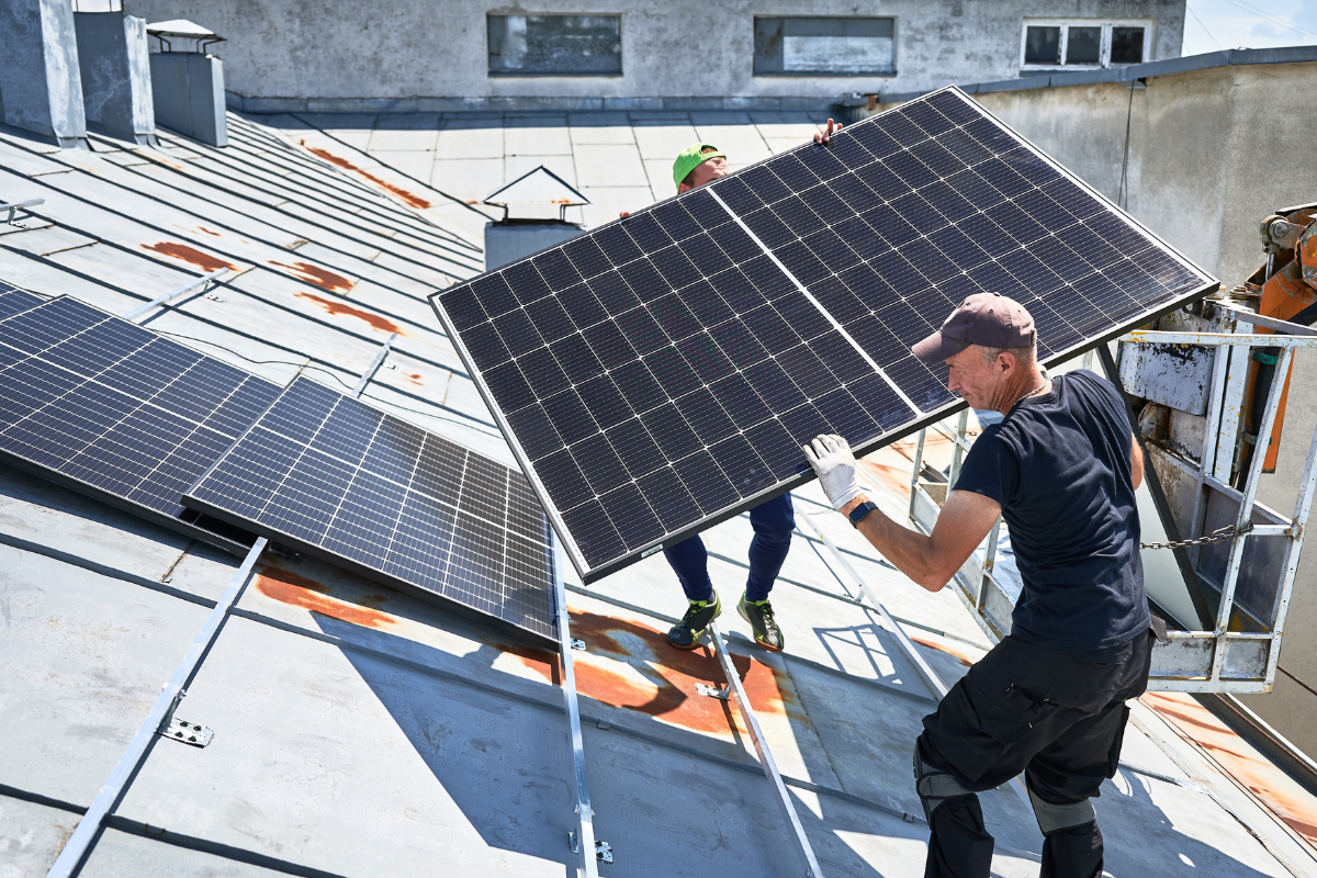Workers building solar panel system on metal rooftop of house with assistance of crane lift. Two men installers carrying photovoltaic solar module outdoors. Renewable energy generation concept