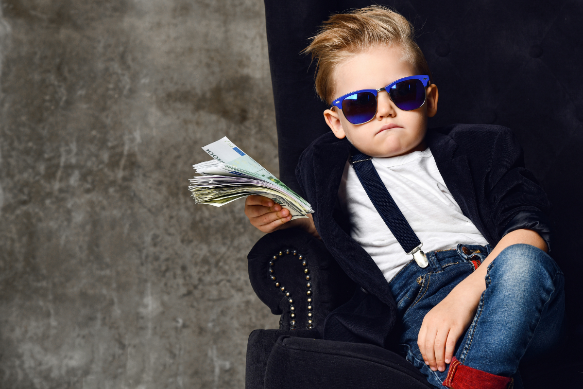 Happy rich kid boy millionaire sits with a bundle of money dollars cash in big luxury armchair