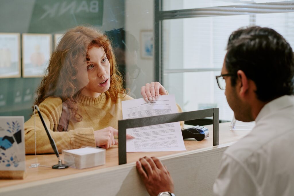 Woman with curly hair discussing financial documents at counter with bank employee focused and engaged in conversation clear glass partition separating them