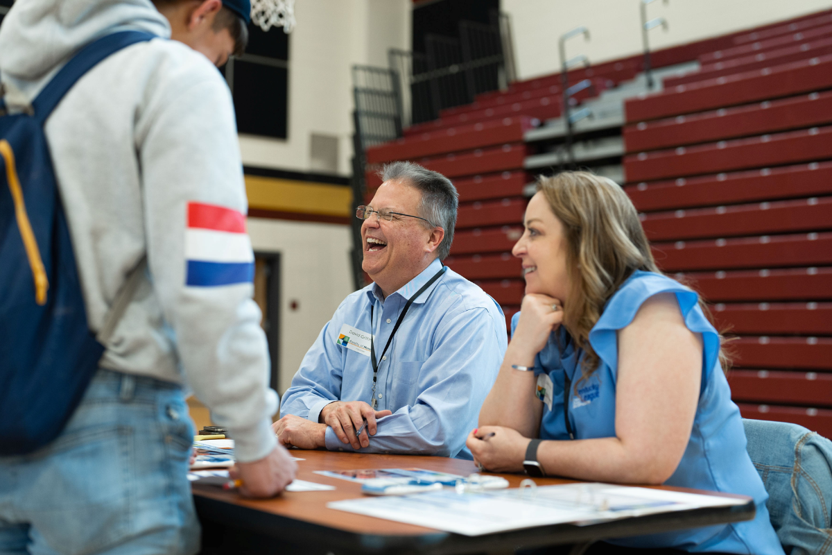Students learn about money management through a real-world simulation at Reality of Money® events, hosted by SECU across the state.with 2 adults sitting at a table and a student standing on the other side looking at information