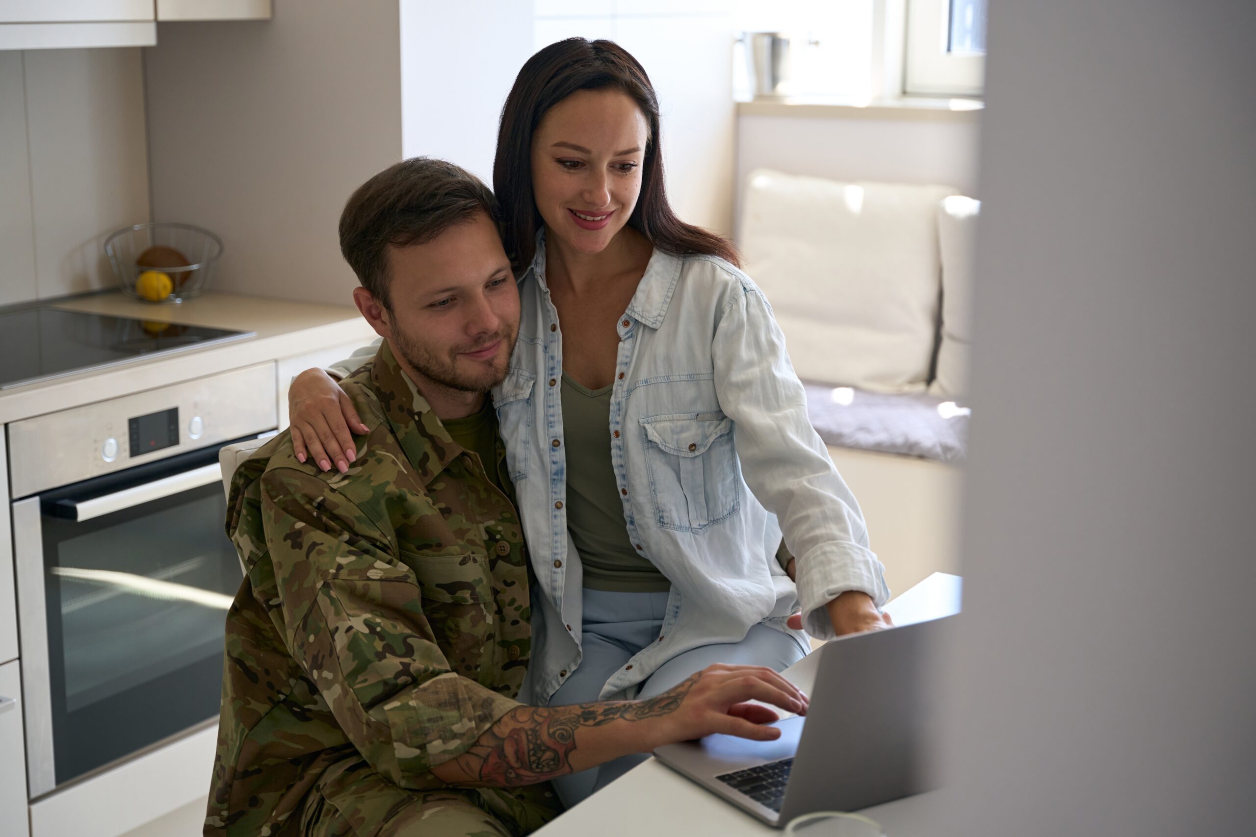 Wife sitting on soldier lap while using laptop in kitchen