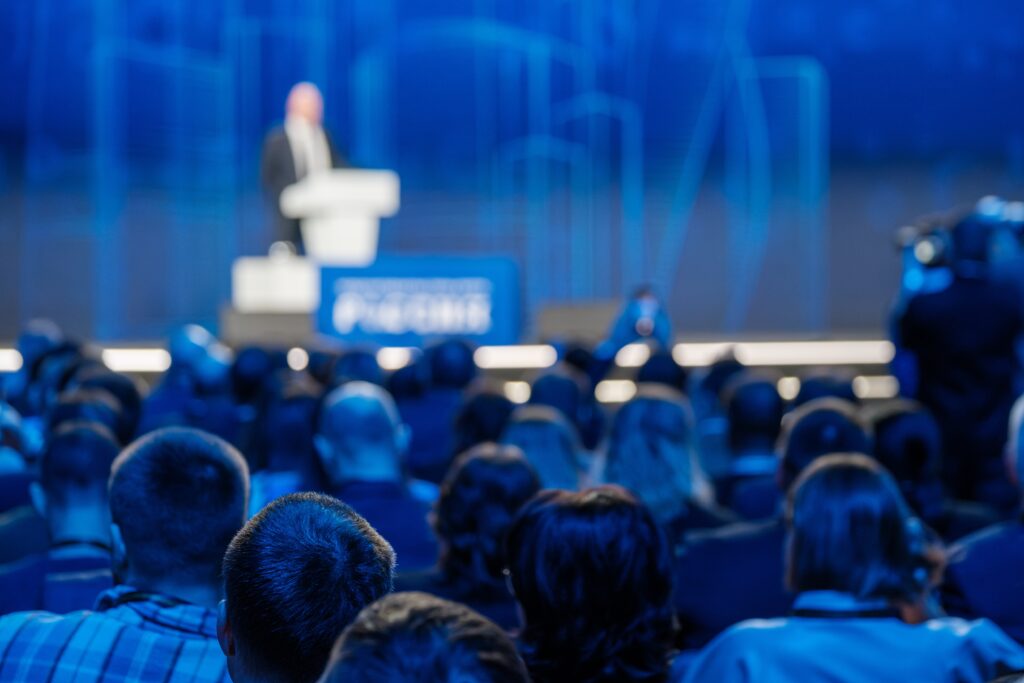 Blurred audience listening to a speaker at a professional conference event in a large auditorium.