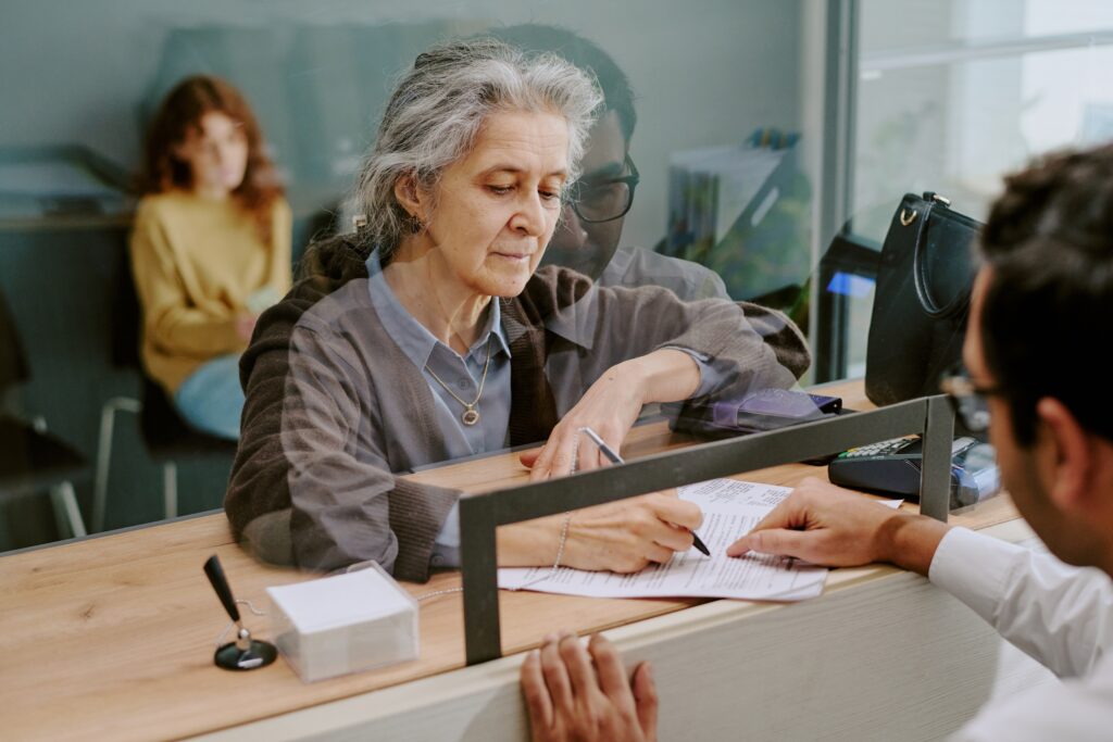 Elderly woman signing official documents at banking counter with teller assisting, seated in modern office setting with reflective glass partition