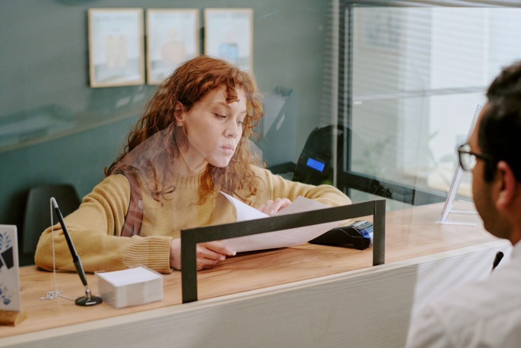 Young woman with curly hair sitting at reception desk while handling paperwork, interacting with unseen individual, including various documents and office supplies for transaction