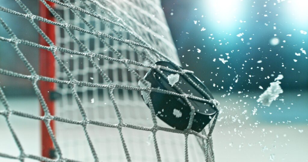 Action sports photo of a hockey puck flying into the goal net during a scoring shot, with ice particles in the air, capturing speed and intensity.