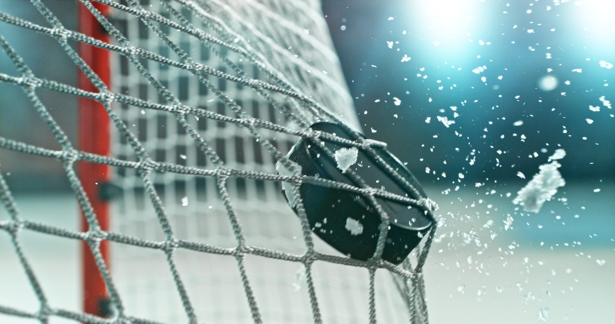 Action sports photo of a hockey puck flying into the goal net during a scoring shot, with ice particles in the air, capturing speed and intensity.