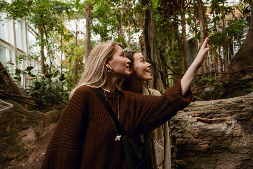 Two White young women with blonde and brown hair are enjoying a moment while exploring nature in a lush indoor forest during their travels, smiling and admiring the surroundings together.