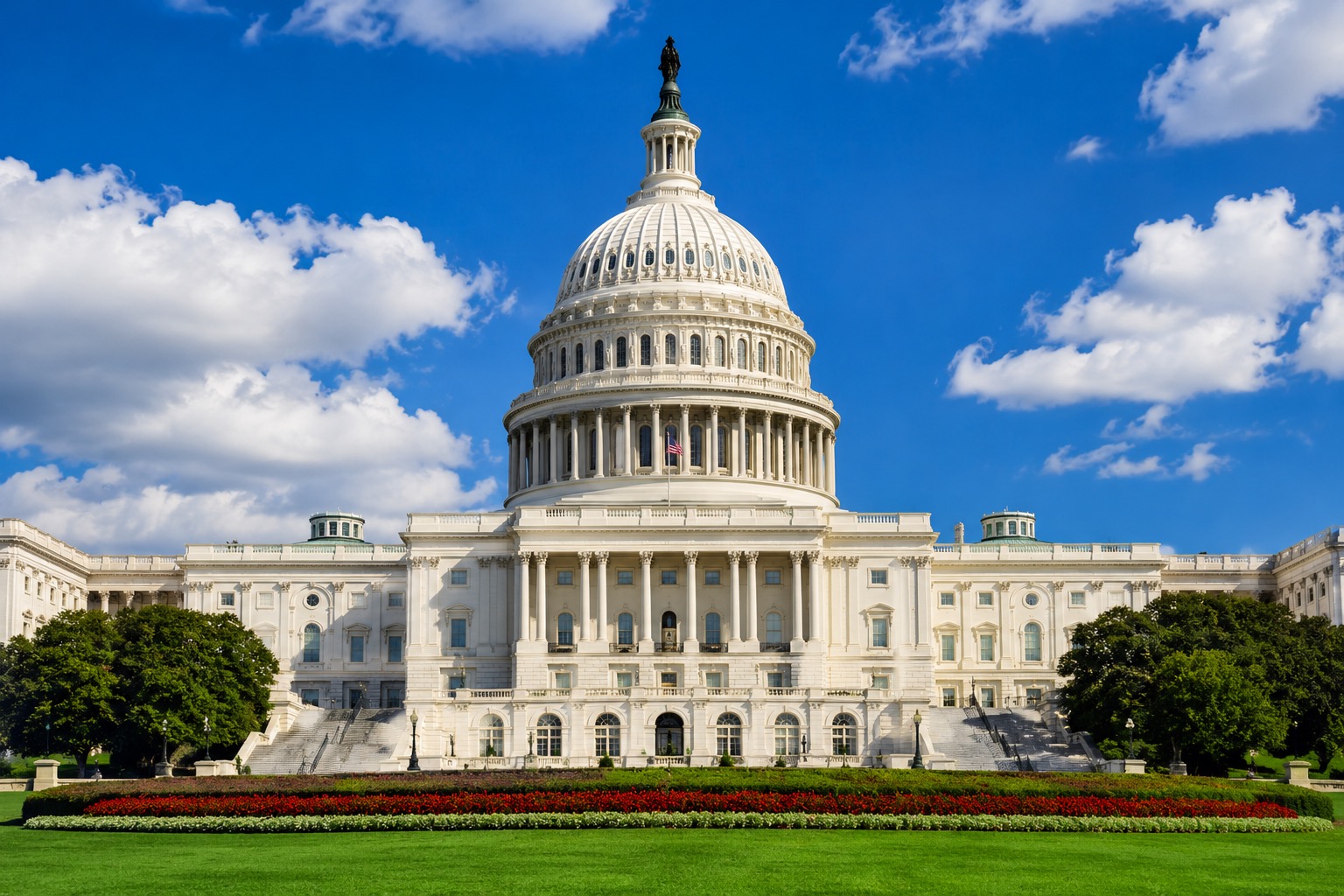 A high-resolution photograph of the United States Capitol in Washington, D.C., captured from the front in perfect symmetry on a bright, sunny day. The iconic white neoclassical building dominates the image, with its massive dome, topped by the Statue of Freedom, rising against a vivid blue sky filled with scattered white clouds. An American flag flies proudly above the east front entrance. The foreground features a lush, manicured green lawn bordered by colorful red flower beds, while mature trees frame both sides of the Capitol. The crisp lighting highlights the building's ornate columns, arches, and architectural details, creating a patriotic and professional image ideal for use in articles or social media posts about Congress, government, legislation, or public policy.
