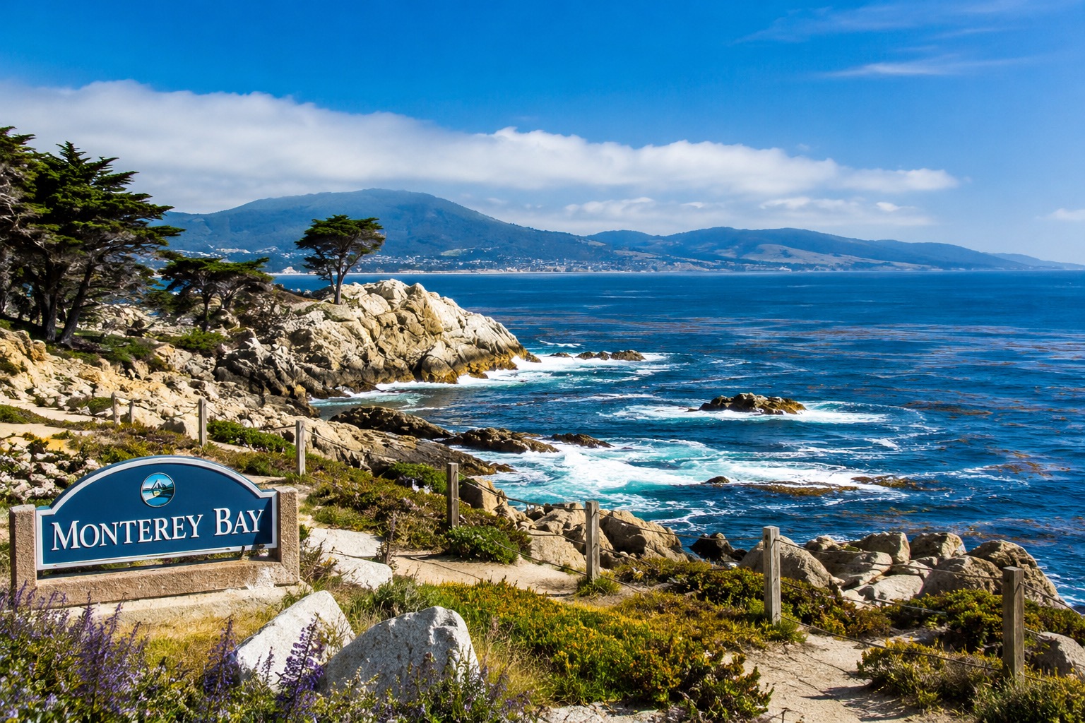 A panoramic coastal view of Monterey Bay, California, showcasing the region's rugged Pacific shoreline under a brilliant blue sky. In the foreground, weathered granite rock formations and windswept Monterey cypress trees overlook the sparkling deep-blue ocean, where white waves crash gently against the rocky coastline. Native coastal vegetation and colorful wildflowers line a scenic walking path, adding vibrant greens and purples to the landscape. The expansive bay stretches toward rolling hills and the distant shoreline, creating a sense of depth and tranquility. A "Monterey Bay" welcome sign in the lower left corner identifies the iconic destination while complementing the scenic composition. The clear weather, crisp visibility, and vivid natural colors capture the beauty of California's central coast, making the image ideal for articles or social media posts about Monterey Bay, marine conservation, tourism, coastal ecosystems, ocean research, or environmental initiatives.