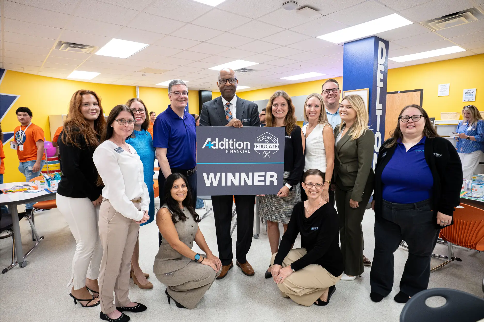 A group of employees from Addition Financial pose together inside a bright, modern classroom after receiving a community recognition award. The group surrounds a large sign that reads "Addition Financial – Renovate to Educate – Winner," proudly celebrating the organization's achievement. The setting features colorful yellow and blue walls, collaborative worktables, and educational materials, suggesting the event took place in a school or learning environment. Several people smile toward the camera, with two team members kneeling in the front while the others stand around the award sign. The central figure, dressed in a business suit, holds the top of the sign as the group gathers closely around. In the background, volunteers and attendees continue participating in activities, reinforcing the atmosphere of a community service or school improvement event. The image conveys teamwork, community engagement, corporate volunteerism, and a shared commitment to supporting education.