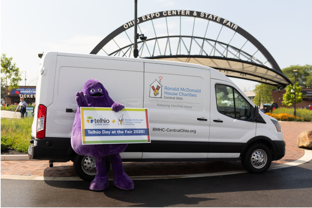 A cheerful community partnership photo taken outside the Ohio Expo Center & State Fair features a purple mascot standing in front of a white Ronald McDonald House Charities of Central Ohio van. The mascot holds a large sign highlighting a partnership between Telhio Credit Union and Ronald McDonald House Charities of Central Ohio for “Telhio Day at the Fair 2026!” The RMHC van prominently displays the organization’s logo along with the message “Keeping families close.” The entrance to the Ohio Expo Center & State Fair is visible in the background, establishing the setting for the event. The image conveys themes of credit union community involvement, charitable partnerships, family support, corporate giving, local engagement, and community-focused events.