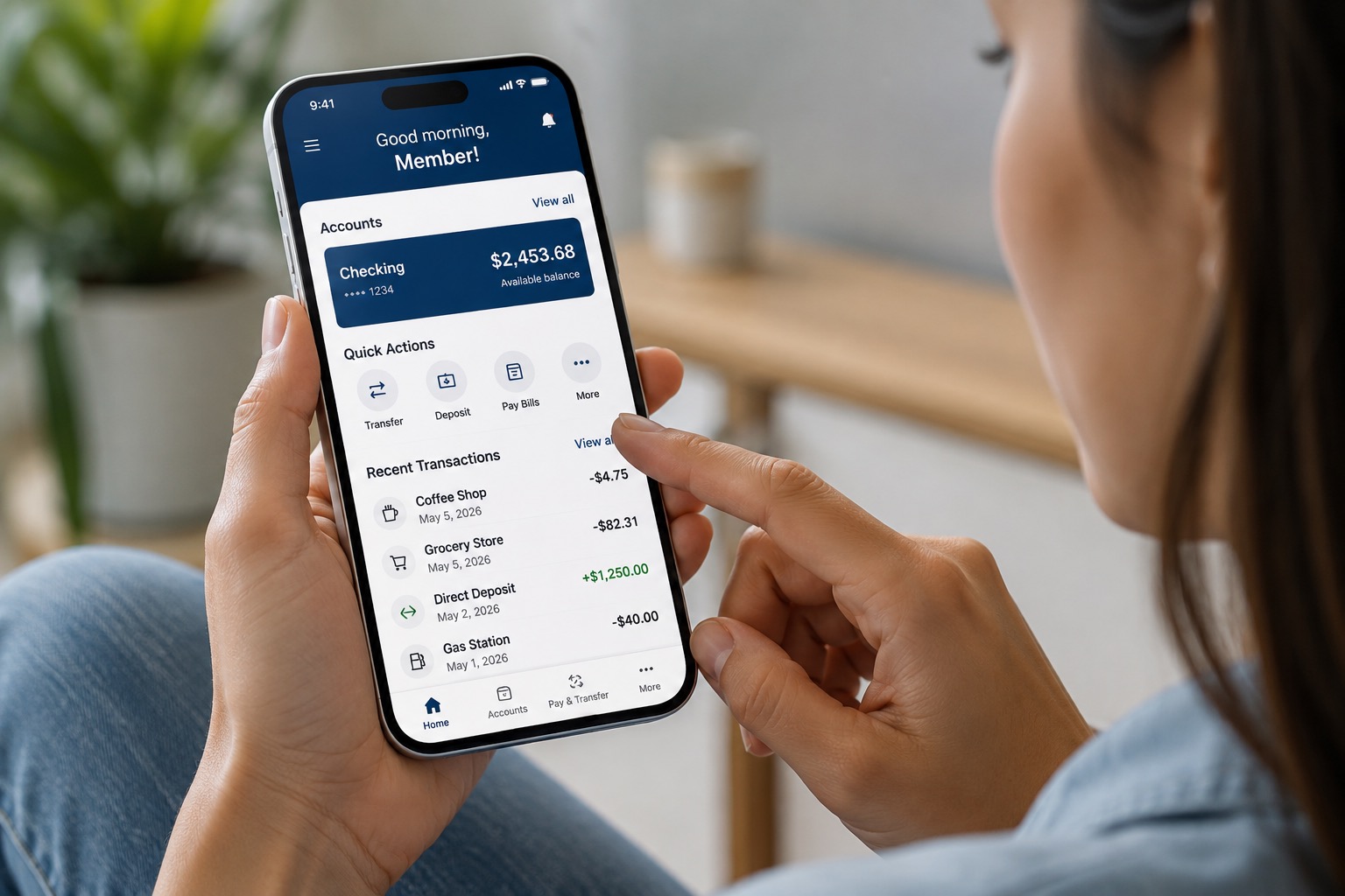 A realistic close-up image shows a person using a digital banking app on a smartphone while relaxing in a comfortable home environment. The phone screen displays a modern mobile banking dashboard with a checking account balance, account information, and recent transactions. Quick-access options allow the user to transfer money, make a deposit, pay bills, and access additional banking services. Recent activity includes everyday purchases, a direct deposit, and other transactions. The person is actively touching the screen, emphasizing the convenience of managing finances directly from a mobile device. The softly blurred background and natural lighting keep the focus on the smartphone and its banking features. The image conveys themes of digital banking, mobile financial services, convenient account access, money management, fintech, and modern credit union technology.