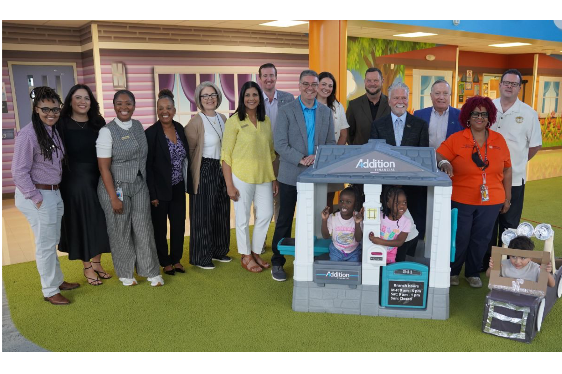 A group photo showing credit union and community leaders gathered with children in a colorful, child-friendly indoor setting. At the center is a miniature playhouse branded Addition Financial, designed to resemble a small credit union branch, with two children inside and another child beside it with a handmade cardboard vehicle. The image highlights themes of community involvement, financial education, youth engagement, partnership, and credit unions investing in the next generation.